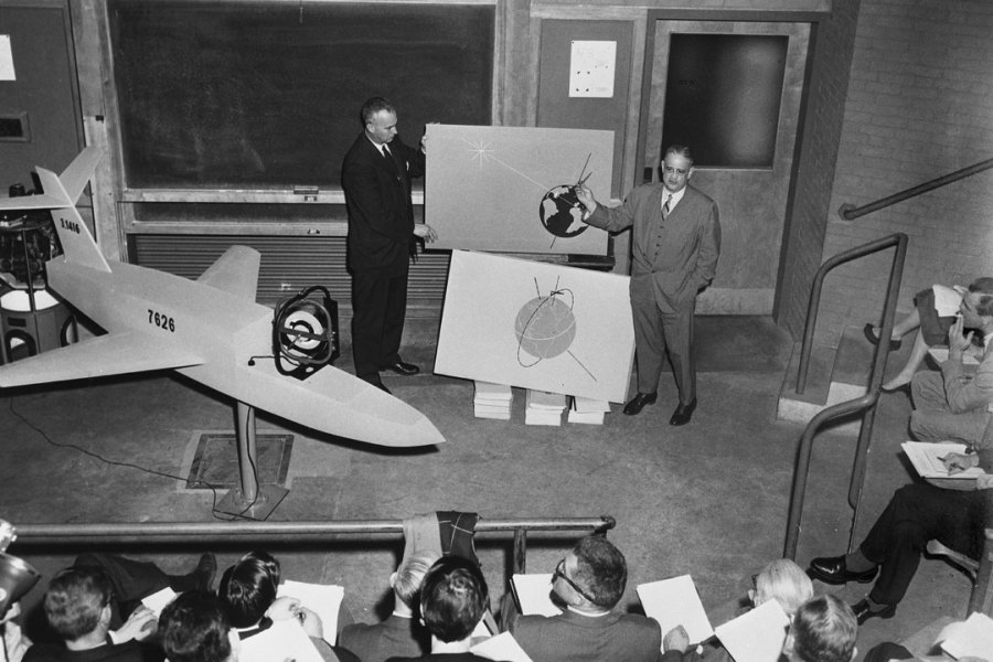 Two men presenting orbital diagrams and an aircraft model to a seated audience in the 1950s